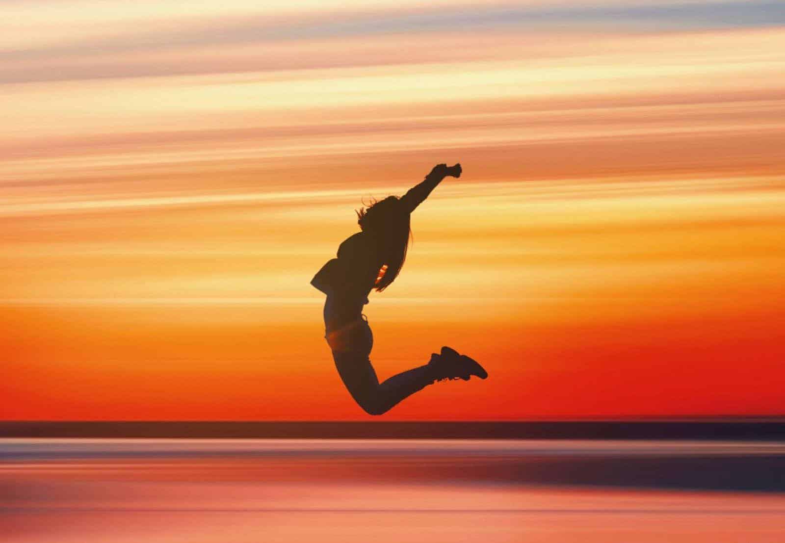 A woman's silhouette jumping up near ocean during bright sunset that fills the entire frame